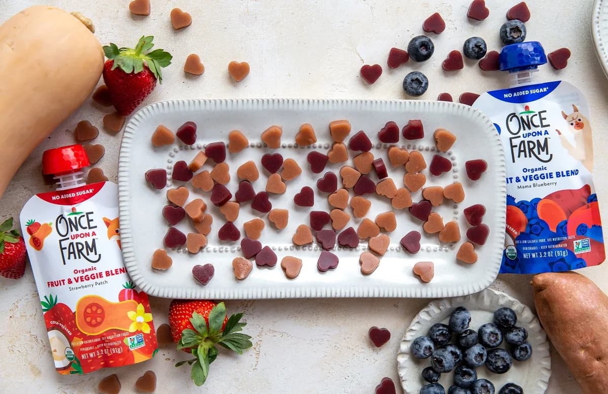 Orange and purple heart-shaped gummies on a white tray beside fruit, vegetables, and Once Upon a Farm pouches